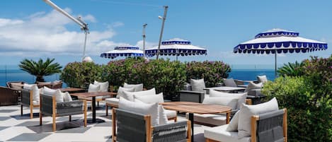 An outdoor seating area with tables and chairs, umbrellas, and a view of the sea.