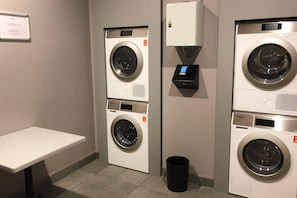 A laundry area with two front-loading washing machines, a small trash bin, and a control panel. - Candlewood Suites Dusseldorf Airport (Düsseldorf)