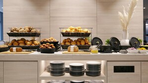 A buffet counter with various breads and pastries, plates stacked on shelves, and kitchen appliances. - Candlewood Suites Dusseldorf Airport (Düsseldorf)