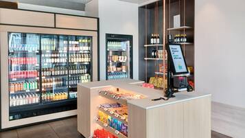 Uma área comercial moderna com expositores refrigerados para bebidas, prateleiras de lanches e um caixa de autoatendimento.