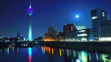 Ein beleuchtetes Stadtbild bei Nacht, mit glänzenden Gebäuden und einem Turm, der sich im Wasser spiegelt.
