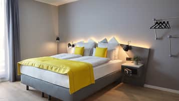 Quarto de hotel moderno com cama, mesa de cabeceira, abajur e um porta-toalhas fixado na parede.