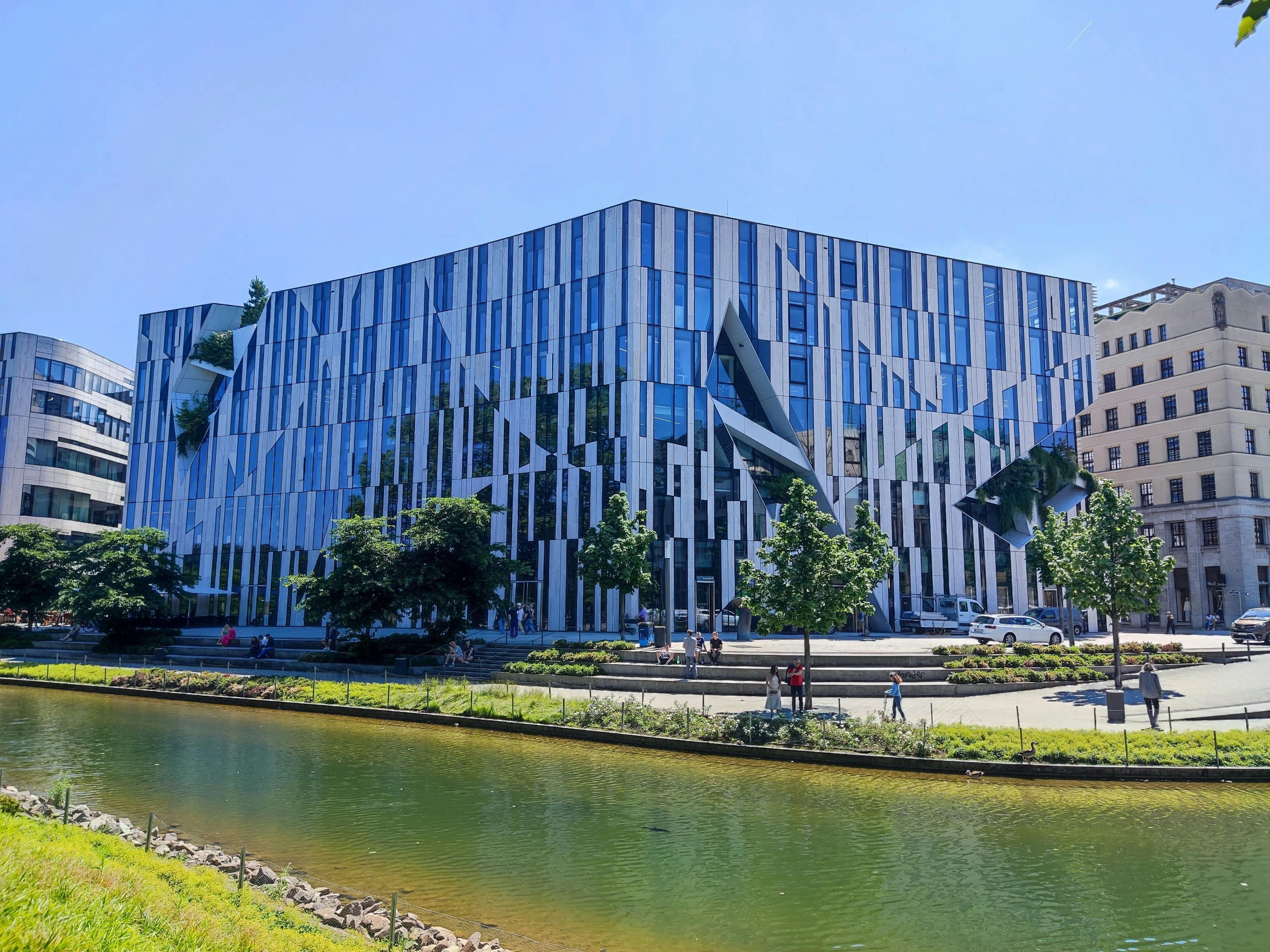 A modern building with a unique geometric facade, a water feature in the foreground, and a clear sky.