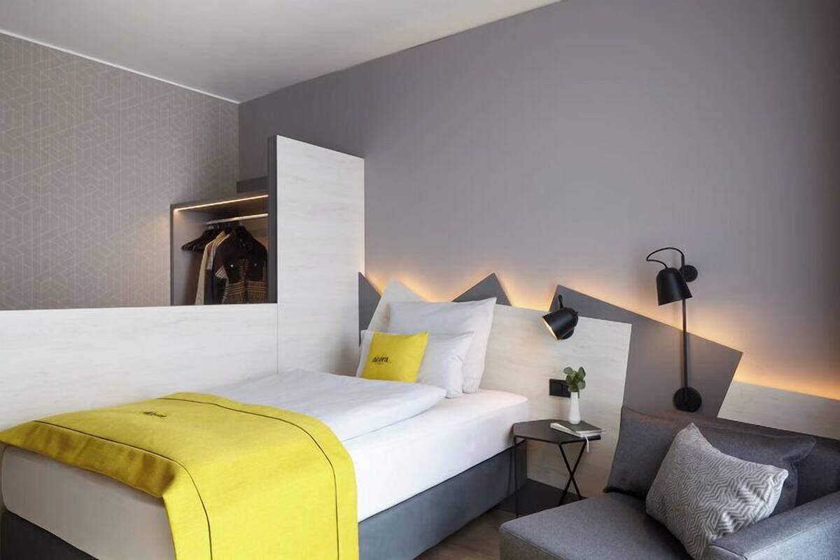 A modern hotel room with a bed, a sofa, a bedside table with a lamp, and a wall-mounted wardrobe.