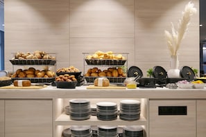 A buffet counter with various breads and pastries, plates stacked on shelves, and kitchen appliances. - Candlewood Suites Dusseldorf Airport (Düsseldorf)