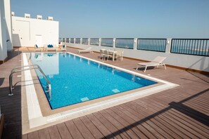 A pool area with a clear view of the sea, a wooden deck, and a white wall with a door. - Shraao Hotel (Doha)