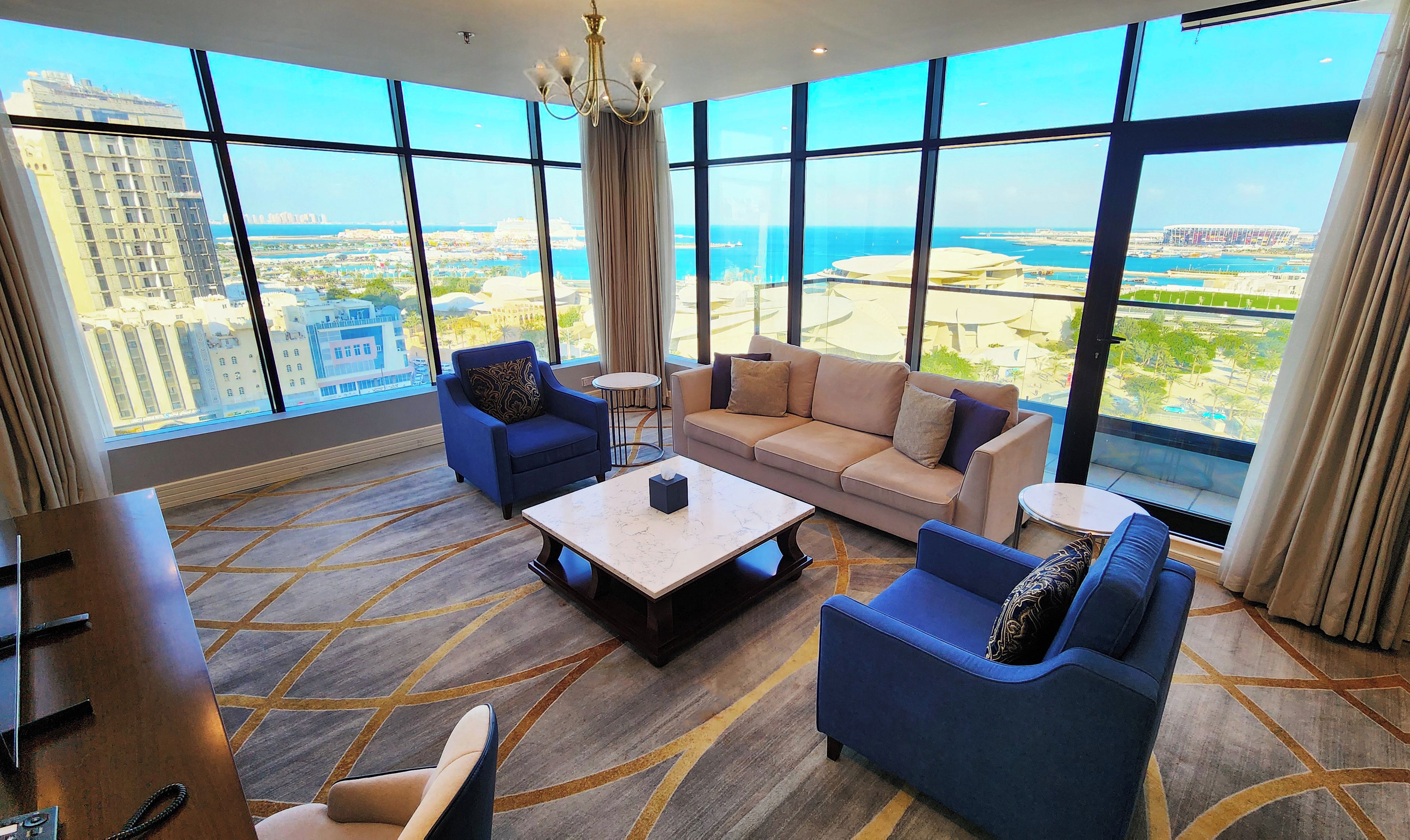 A hotel room with a large window, a sofa, a chair, a coffee table, and a desk.
