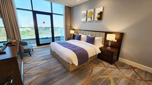 A hotel room with a large bed, a desk, a chair, a nightstand, and a view of the city through large windows. - Shraao Hotel (Doha)