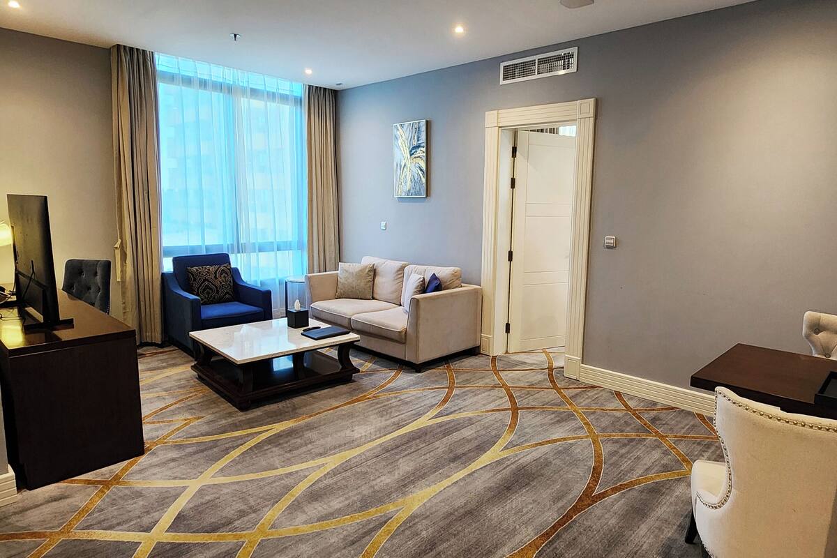 A modern hotel room with a sofa, armchair, and a small table. There is a large window with curtains, a painting on the wall, and a door.