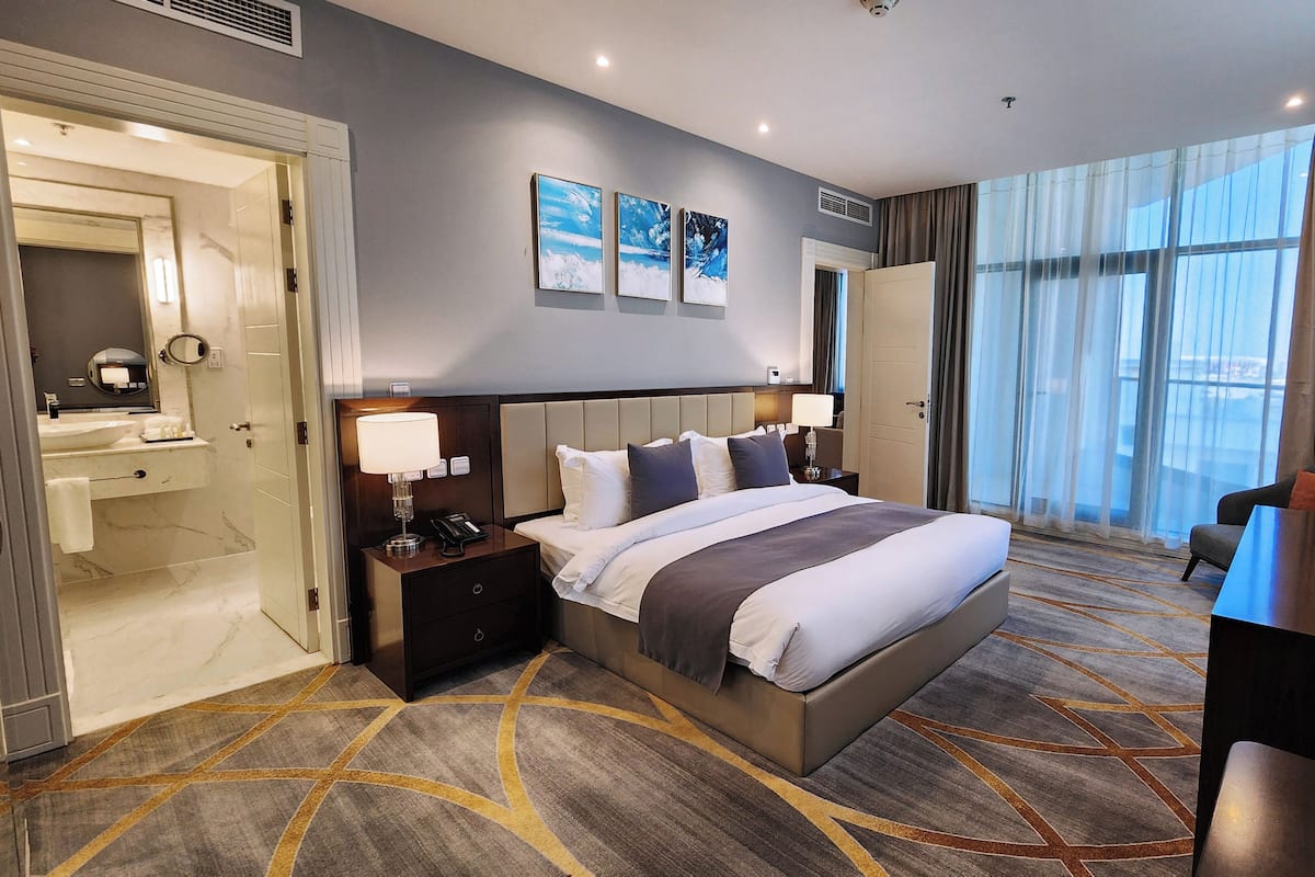 A hotel room with a large bed, bedside tables, a desk, a chair, a bathroom with a mirror, and artwork on the wall.