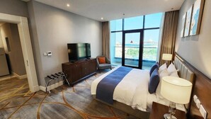 Une chambre d’hôtel avec un grand lit, une télévision, une chaise, un bureau avec une lampe et une vue sur la ville grâce à de grandes fenêtres.