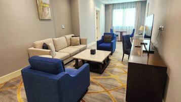 Quarto de hotel com sofá, duas poltronas, mesa de centro, televisão e escrivaninha.