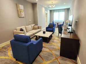 Quarto de hotel com sofá, duas poltronas, mesa de centro, televisão e escrivaninha.