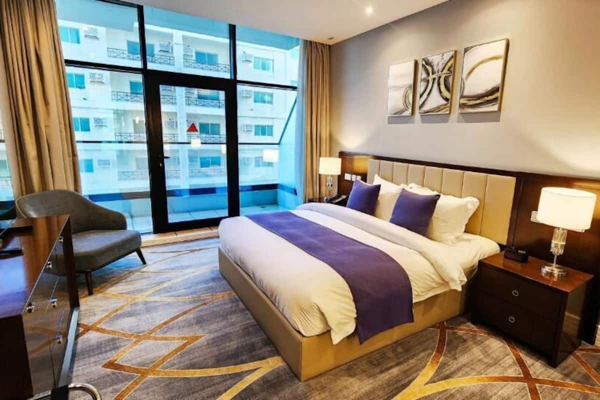 A hotel room with a large bed, a desk, a chair, and a view of the city through large windows.