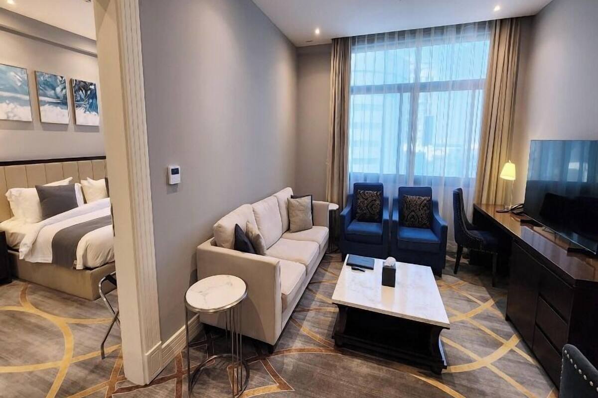 A hotel room with a bed, a sofa, two armchairs, a coffee table, a TV, and a large window with curtains.