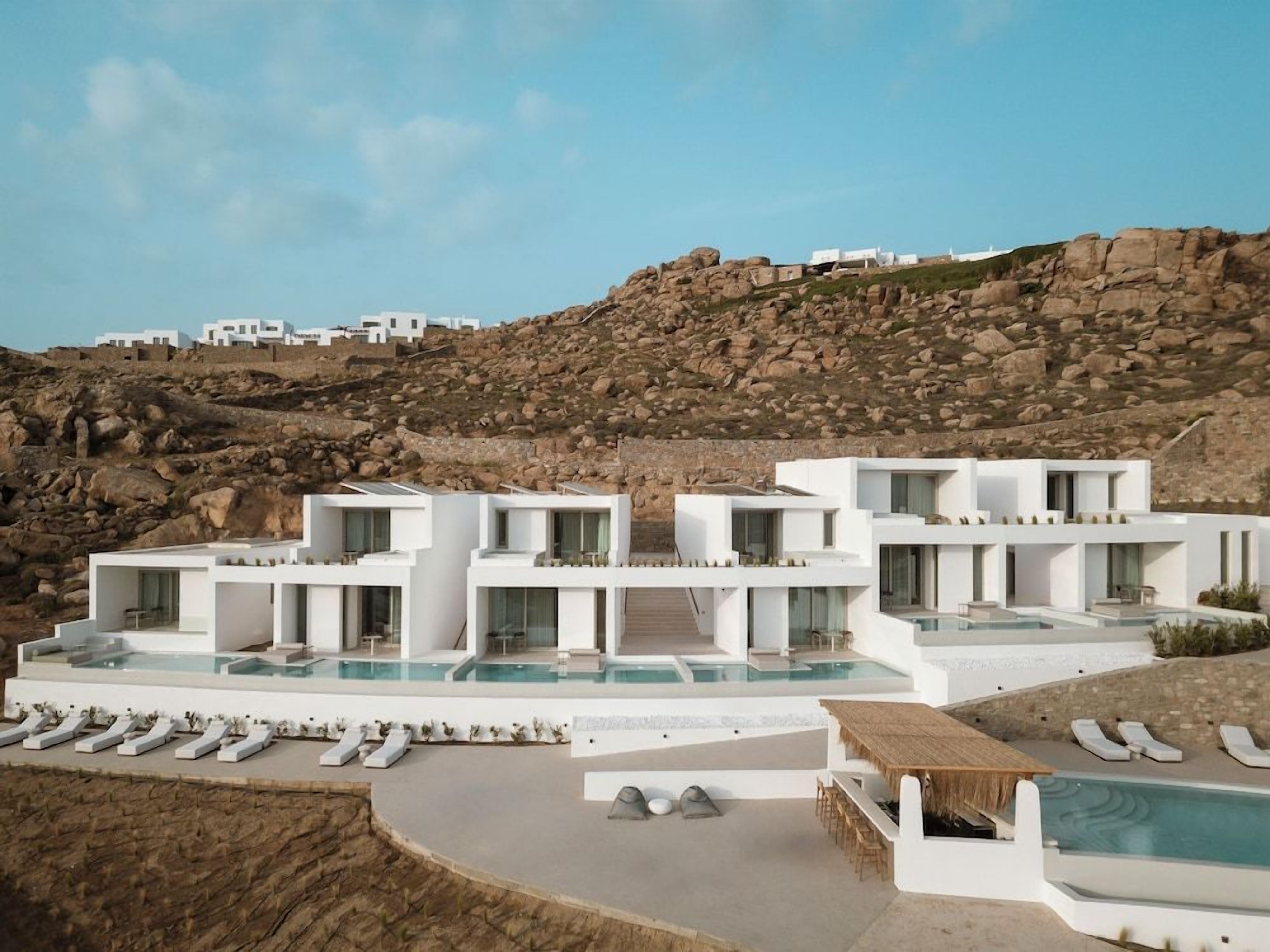 A modern resort with multiple white buildings, a pool area, and a rocky hillside backdrop.