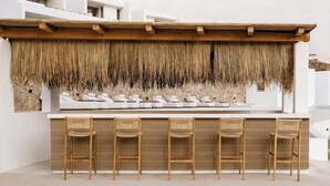Zona de bar amb sostre de fusta, paret de palla i tamborets blancs.