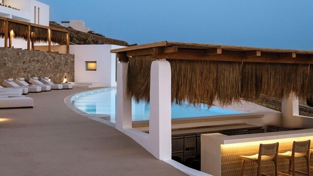 Zona de piscina amb gandules, bar i una estructura amb teulada de palla.