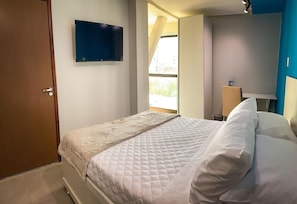 Une chambre à coucher avec un lit, un téléviseur fixé au mur et une fenêtre laissant entrer la lumière naturelle.
