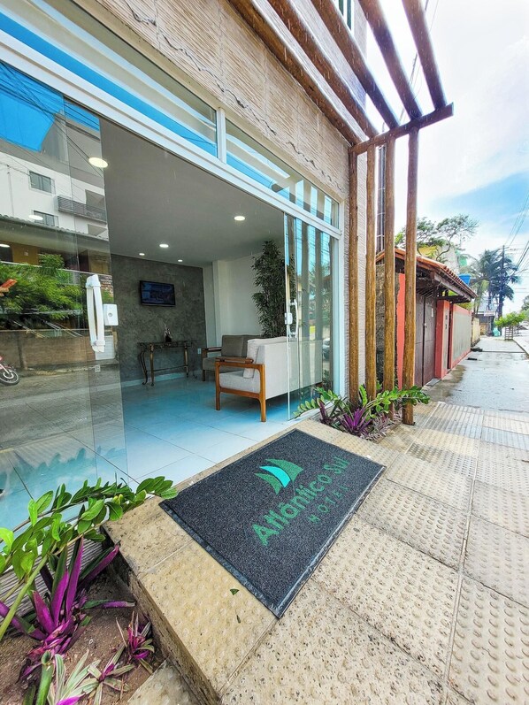 Una entrada de hotel moderna con fachada de vidrio, un área de recepción con sofá y sillones, y un felpudo con el nombre del hotel.