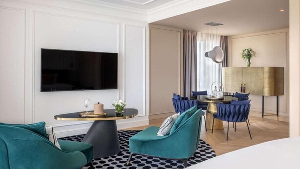 Een moderne woonkamer met een flatscreen televisie, smaragdgroene fauteuils, een ronde salontafel en een eethoek met blauwe stoelen en een gouden lamp.
