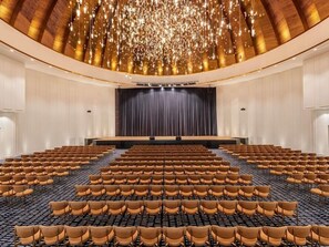Een grote zaal met rijen stoelen, een podium met gordijn en een plafond met hangende lampen.