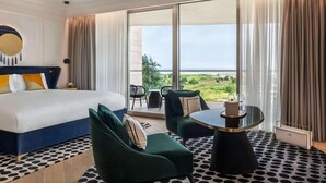 Habitación de hotel con una cama grande, dos sillones verdes, una mesita redonda y un balcón con vistas al mar.