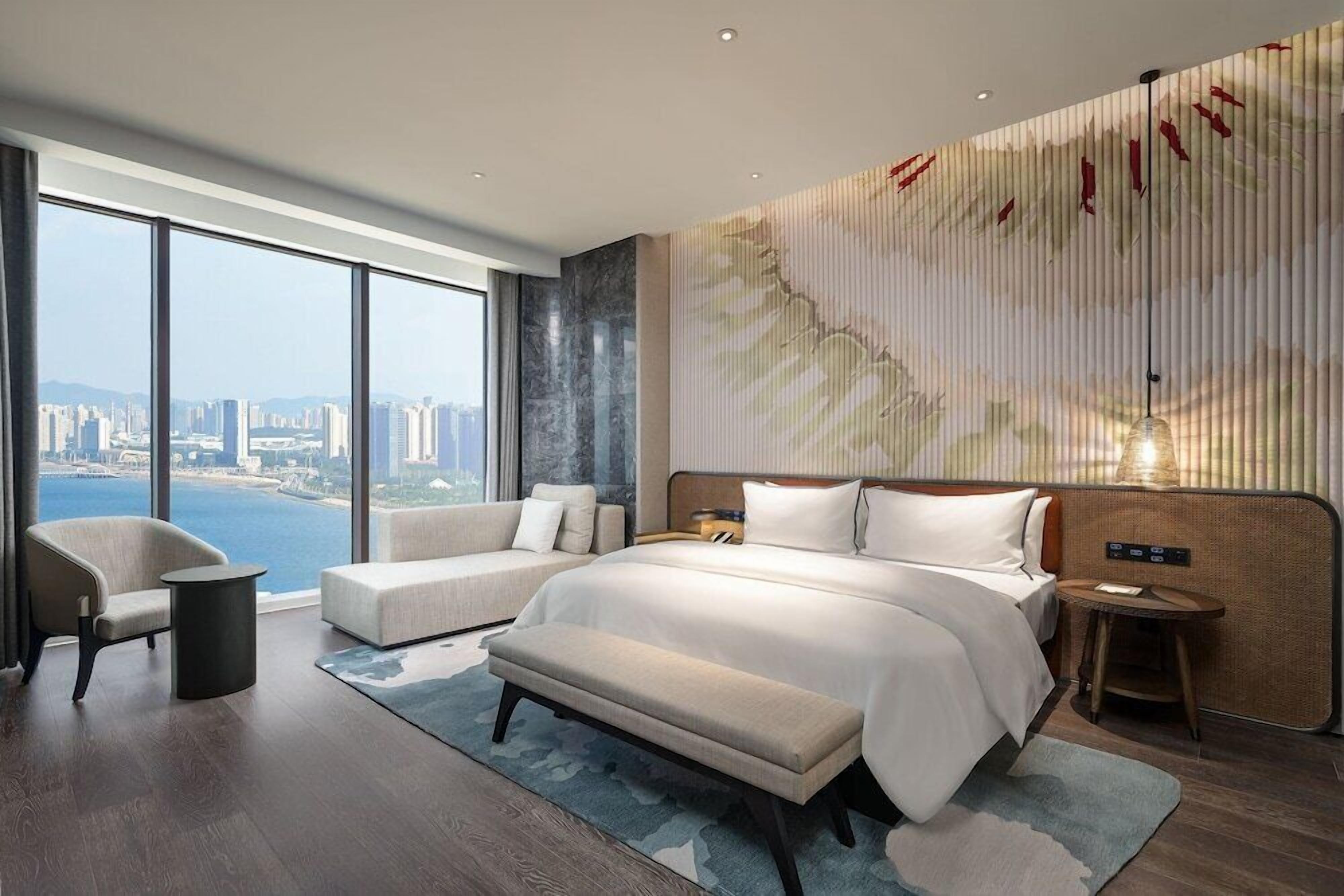 A modern hotel room with a large bed, a sofa, a bench, and a view of a cityscape through large windows.