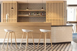 Uma área de bar com balcão de madeira, três taburetes brancos e prateleiras com garrafas e copos.