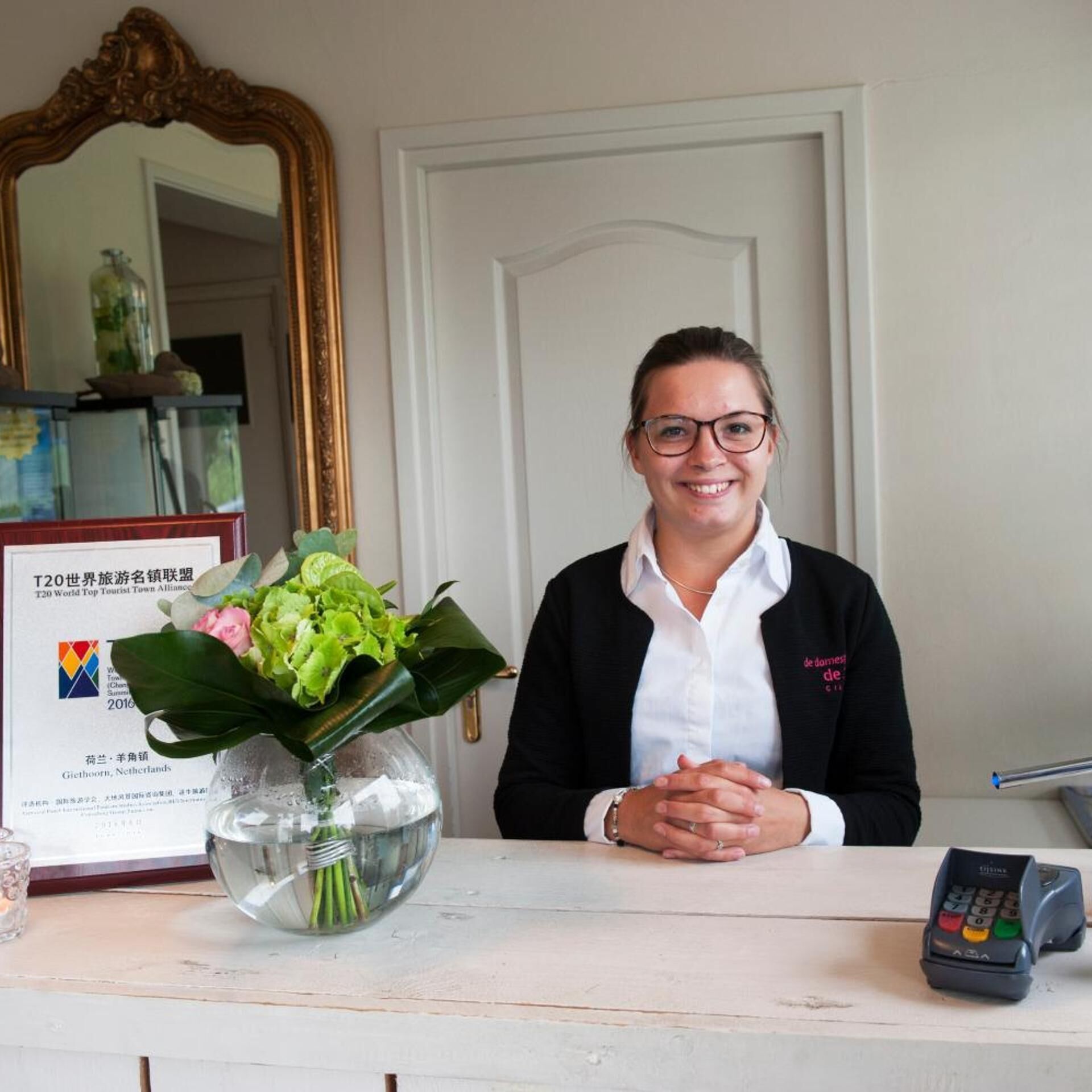 A reception desk with a framed certificate, a vase with flowers, and a payment terminal.