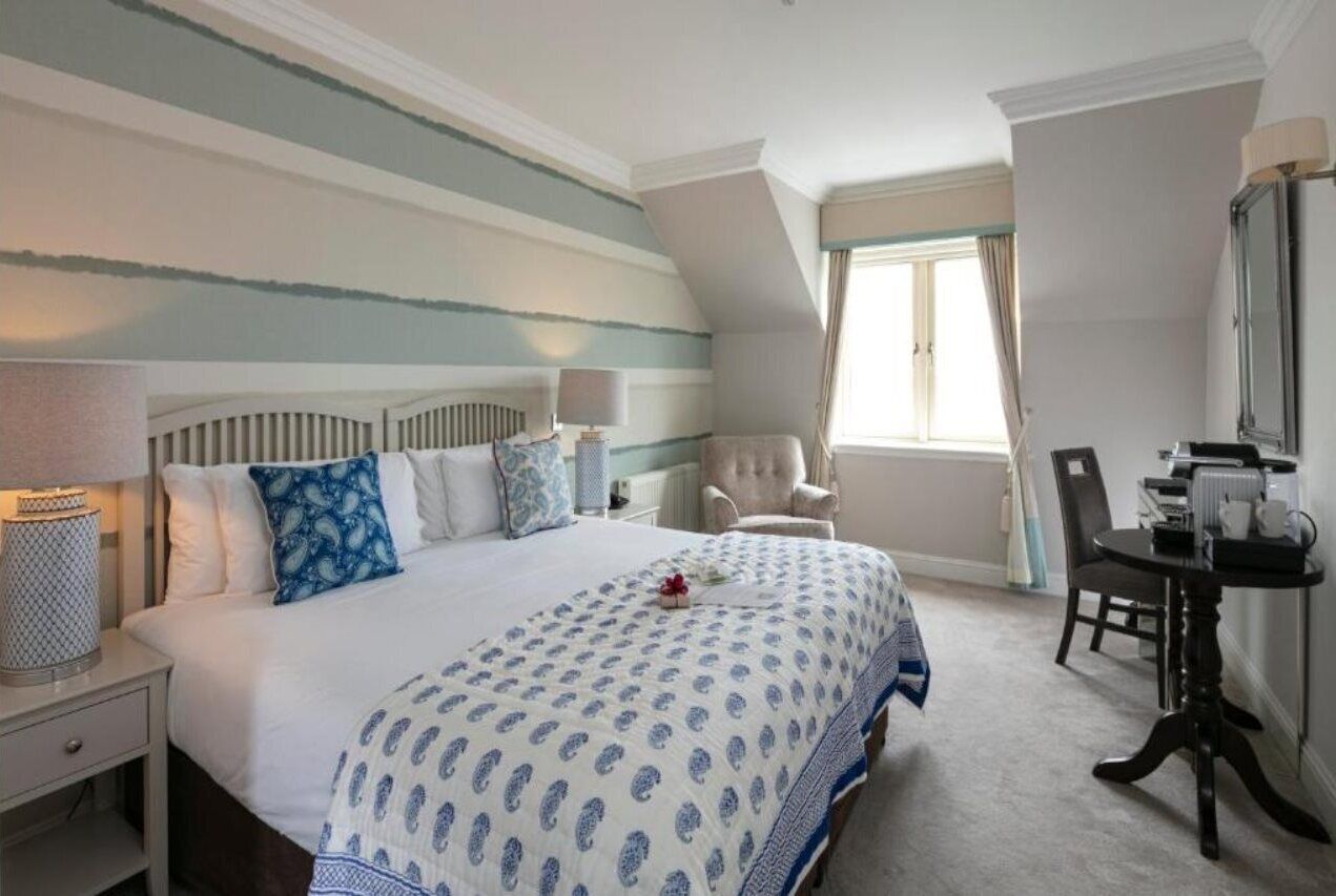 A hotel room with a large bed, two bedside tables, a chair, a small table, and a window with curtains.