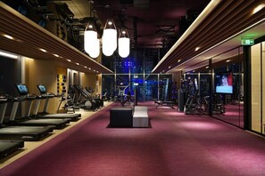 Uma academia com esteiras, máquinas de musculação e piso revestido de carpete vermelho.