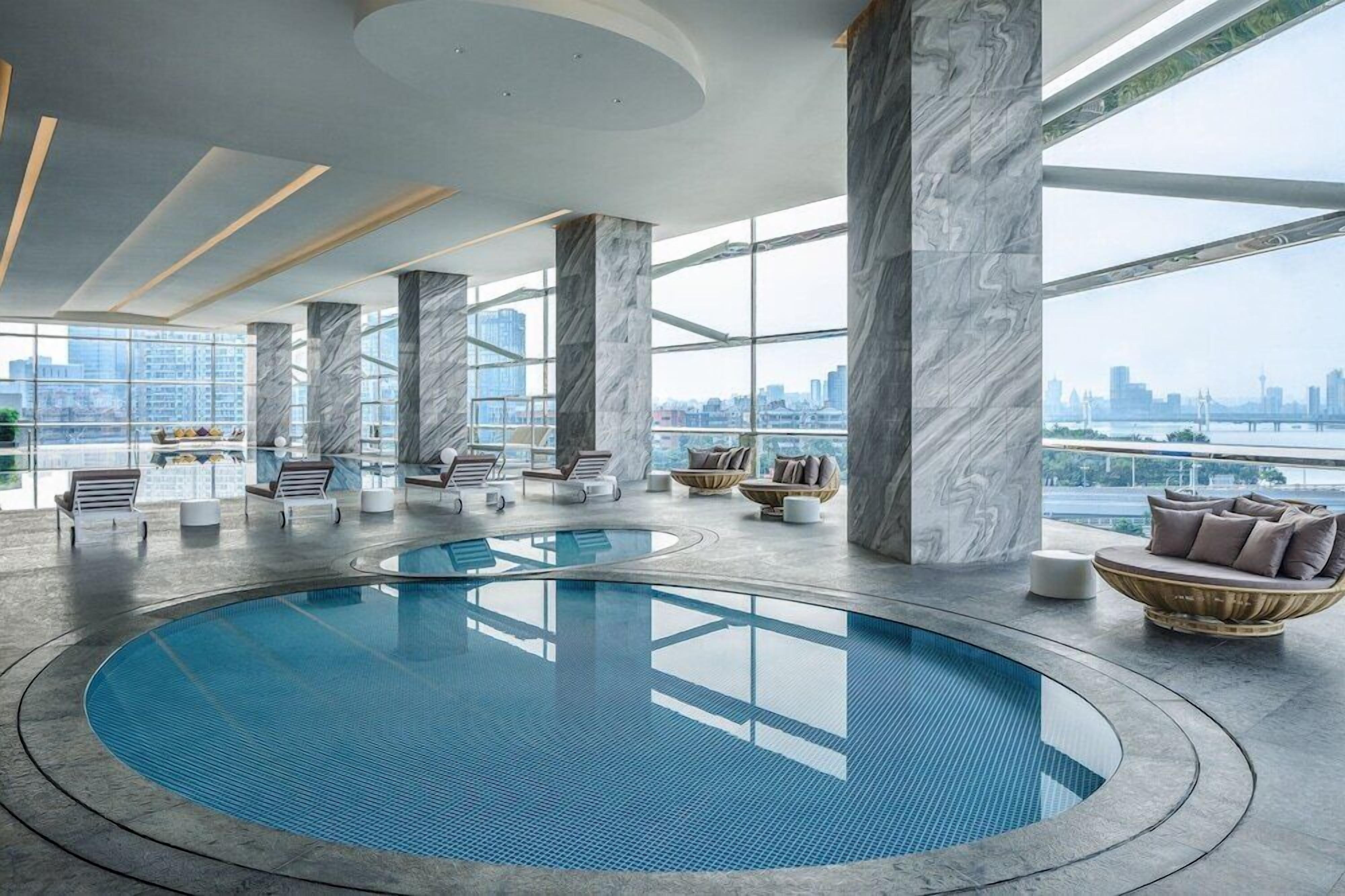 A modern indoor pool with marble columns, lounge chairs, and a city view.