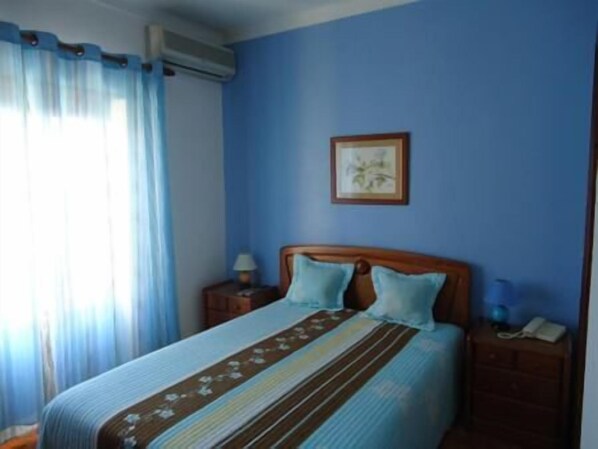 A bedroom with a bed, a nightstand, a lamp, a framed picture on the wall, and a window with curtains. - Hotel Conforto Latino (Abrantes)