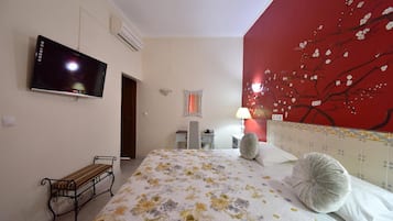A bedroom with a bed, a television mounted on the wall, a bench, a lamp, and a red wall with a tree painting.