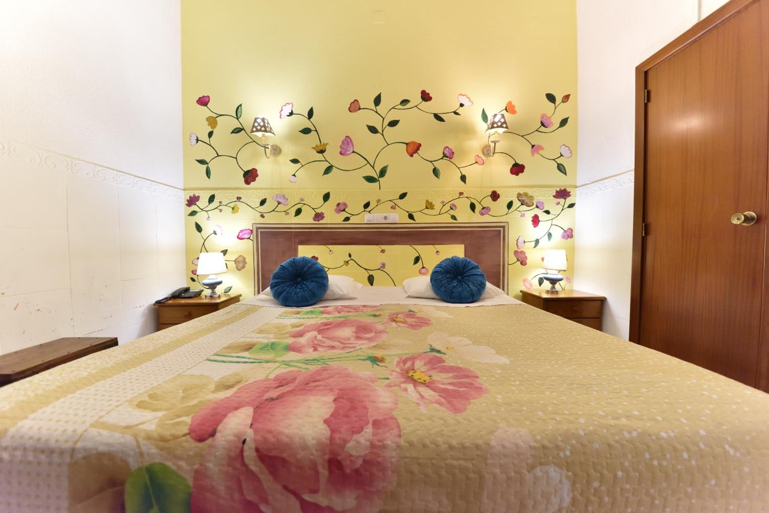 A hotel room with a floral-patterned bedspread, a wooden headboard, and wall-mounted lamps.