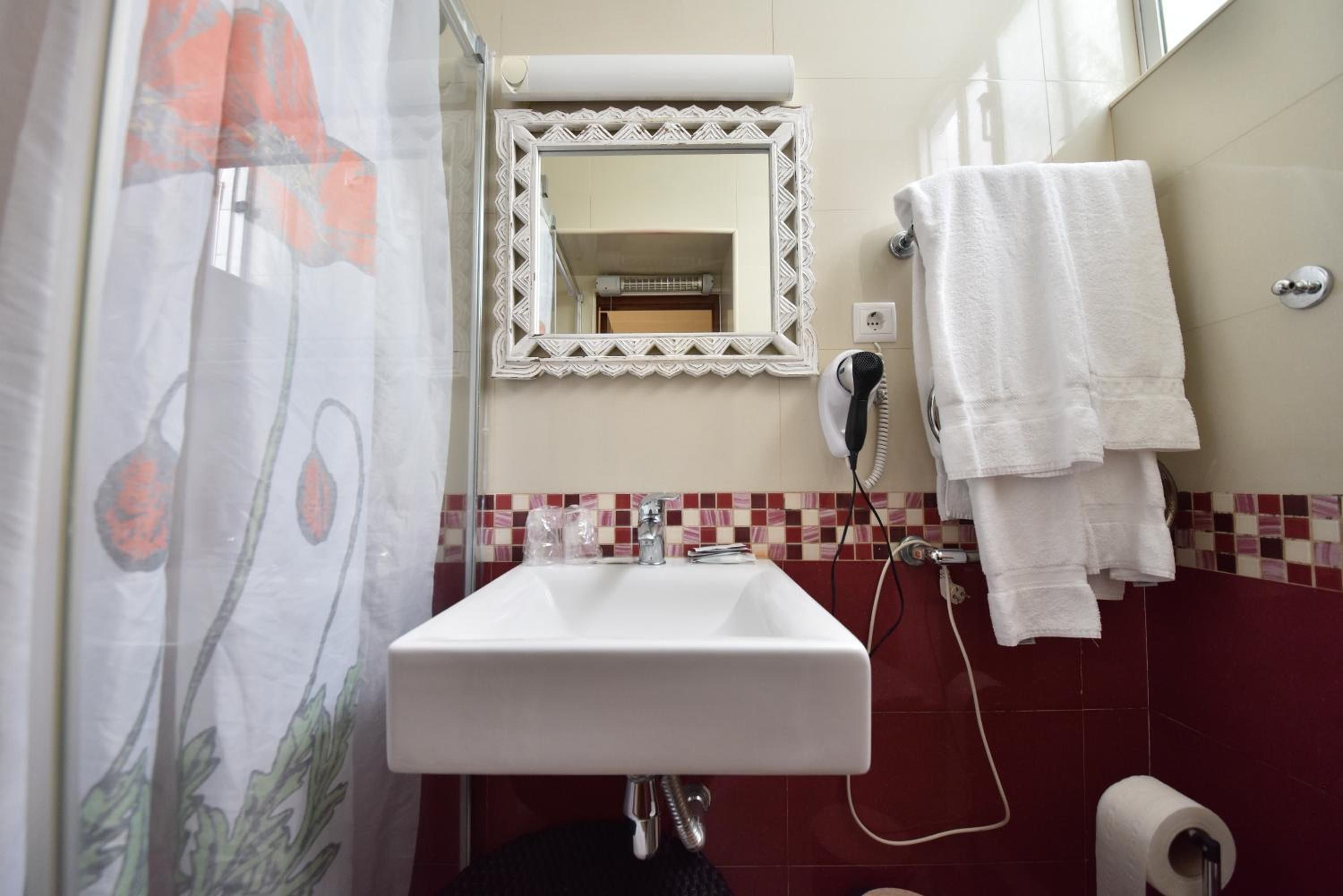 A bathroom with a white sink, a mirror with a decorative frame, a towel rack with towels, a shower curtain with a floral pattern, and a tiled wall with a red and beige mosaic backsplash.