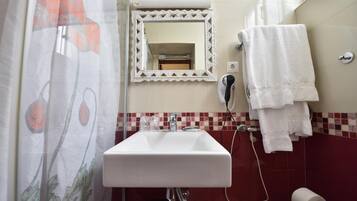 A bathroom with a white sink, a mirror with a decorative frame, a towel rack with towels, a shower curtain with a floral pattern, and a tiled wall with a red and beige mosaic backsplash.