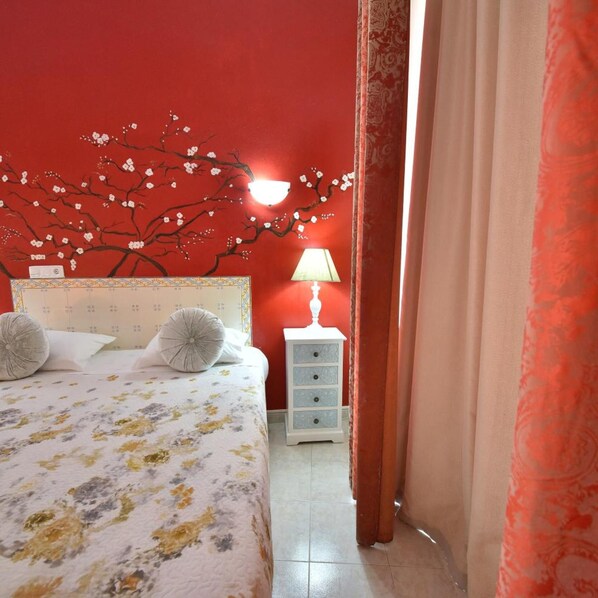 A bedroom with a red wall, a bed with floral bedding, a nightstand with a lamp, and a calendar on the wall. - Hotel Bejense (Beja)