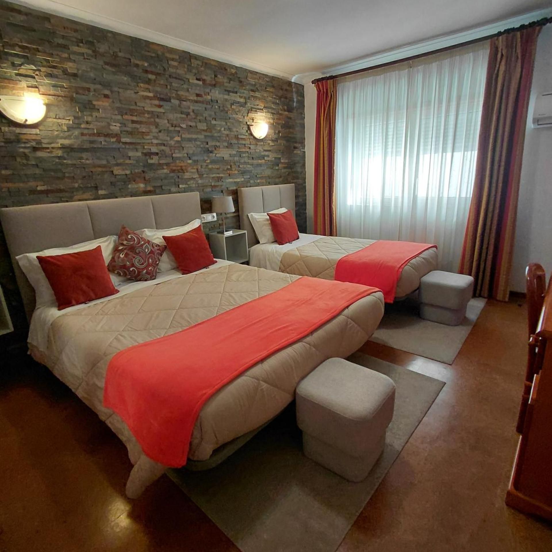 A hotel room with a stone wall, two beds with red bedding, a nightstand, and a window with curtains.