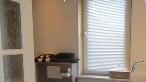 A bathroom with a toilet, a sink, a towel dispenser, and a window with blinds. - Hotel Hoogland Zandvoort Aan Zee (Zandvoort)