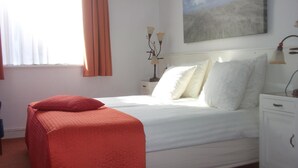 A neatly made bed with white linens and a red throw blanket, a bedside table with a lamp, and a painting on the wall. - Hotel Hoogland Zandvoort Aan Zee (Zandvoort)