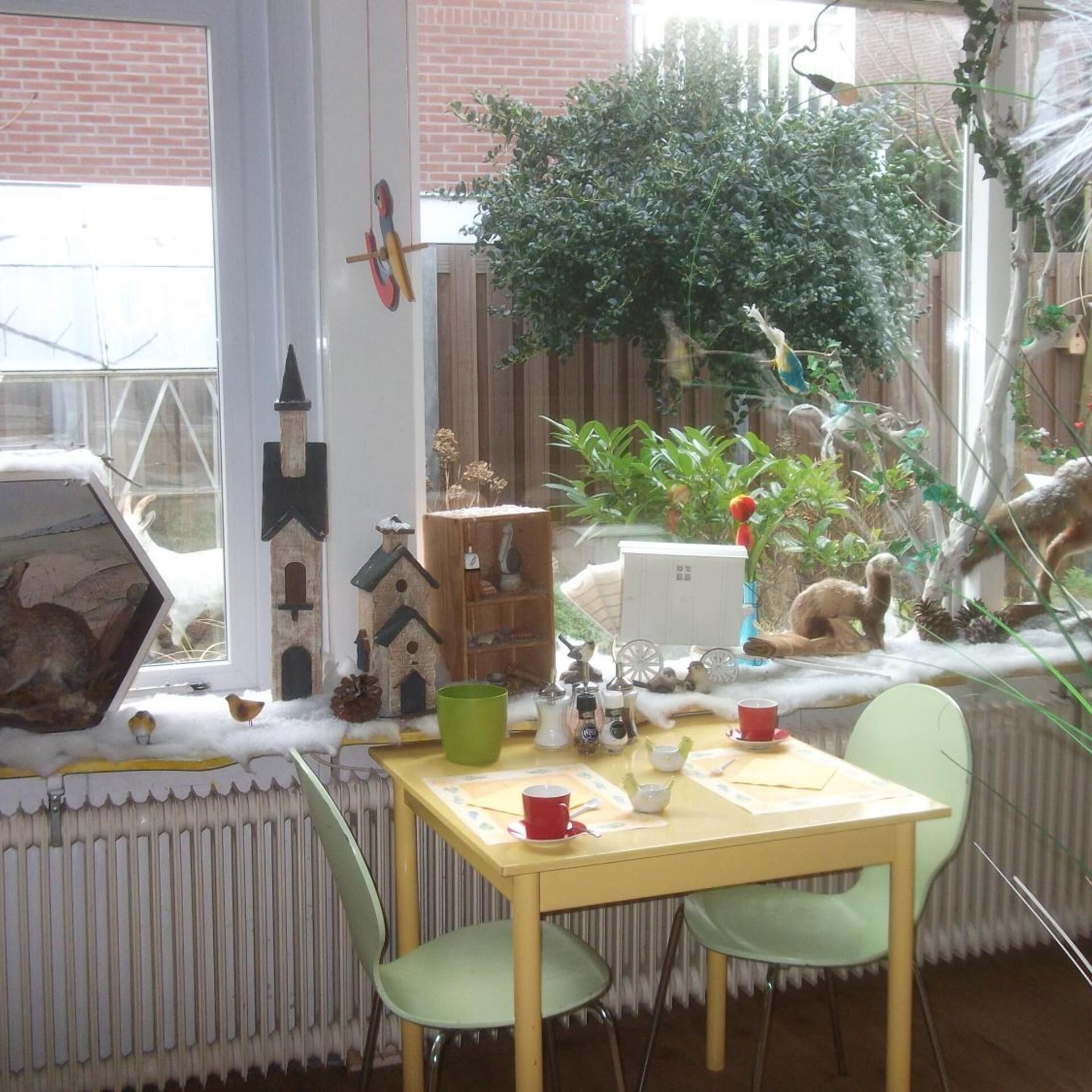 Een kamer met een tafel, stoelen, een radiator, een raam en uitzicht op een tuin.