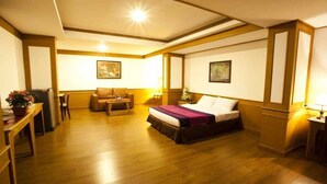 A hotel room with a wooden floor, a bed with a purple bedspread, a desk with a lamp, a chair, and a small table with a plant. - Zaza Hotel (Samut Prakan)