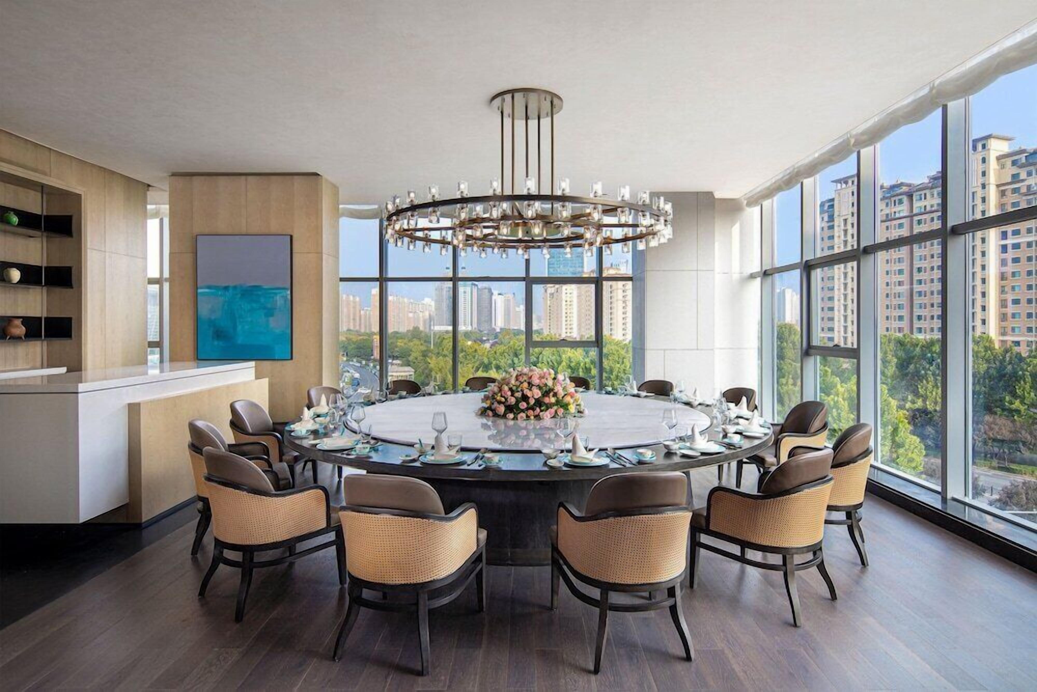 A modern dining room with a round table, wicker chairs, a chandelier, and a city view through large windows.