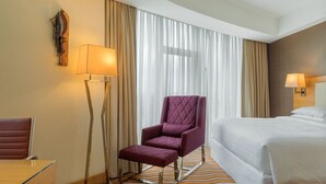 Ett hotellrum med en säng, en stol, ett skrivbord, en lampa och gardiner.