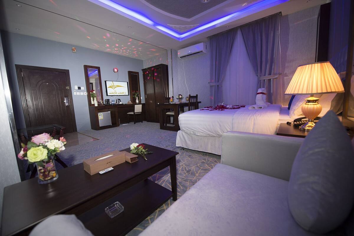 A hotel room with a bed, a sofa, a desk, a TV, and an air conditioning unit.