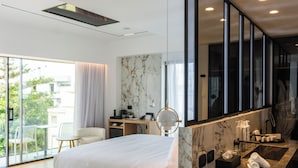 A modern hotel room with a large bed, a sitting area with a desk, a bathroom with a large mirror, and a balcony with a view of the city. - Ethereal White Resort Hotel & Spa Adults Only 16+ (Heraklion)