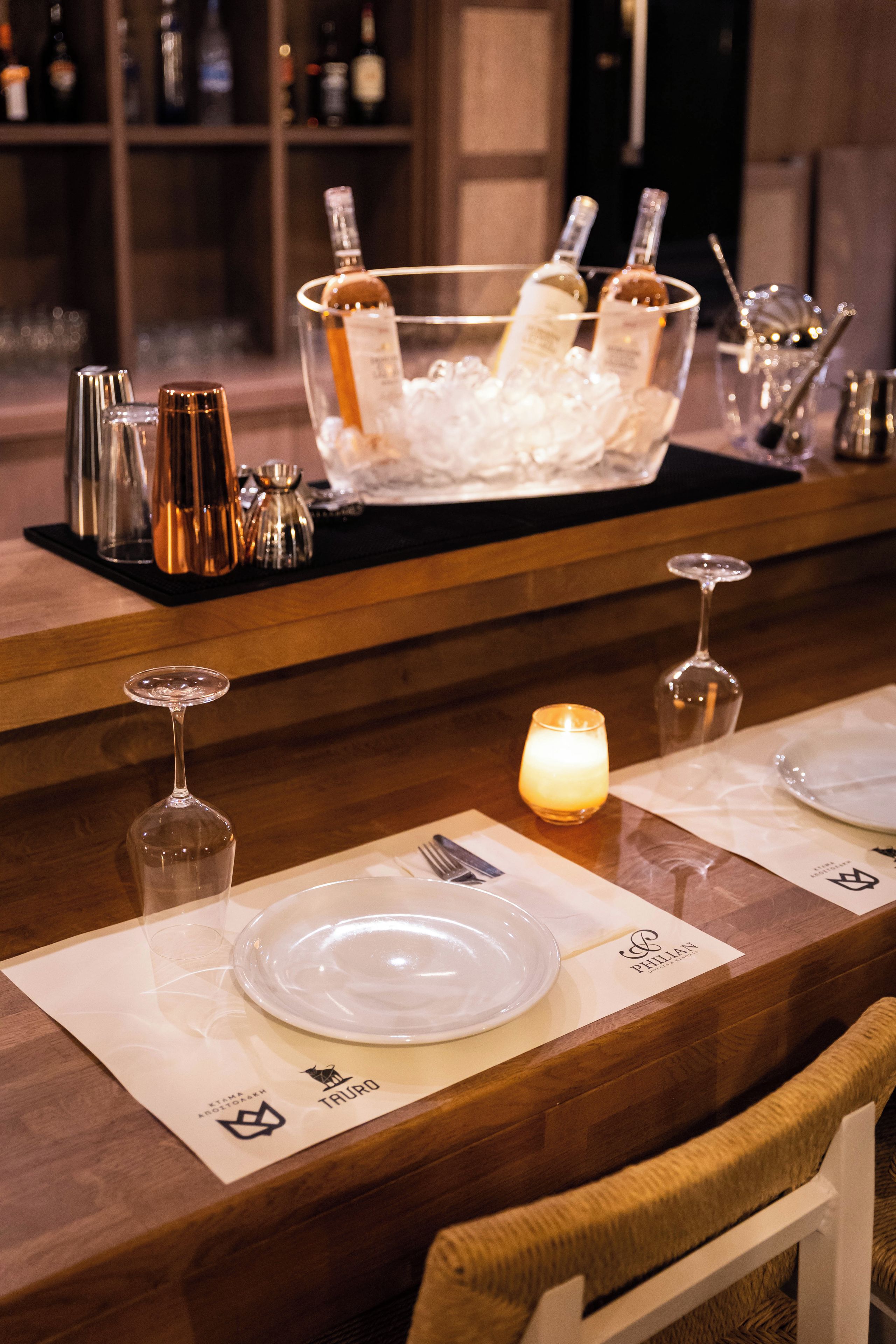 Un espace bar avec un comptoir en bois, un récipient en verre contenant des bouteilles, des verres et une bougie.