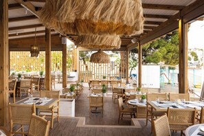 An outdoor dining area with wooden tables and chairs, potted plants, and a thatched ceiling. - Skiathos Theros (Skiathos)